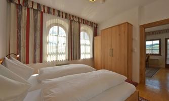 A cozy bedroom with two beds and a wooden wardrobe. Large windows with curtains provide plenty of daylight.