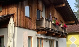 A traditional wooden house with a balcony full of flowers. The garden is surrounded by a green hedge.