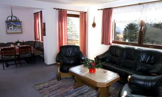 A cozy living room with black leather furniture and a wooden table. Large windows let in plenty of light and provide a view of the outdoors.
