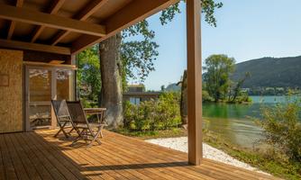 A cozy terrace with a wooden floor and two loungers overlooks a tranquil lake. Surrounded by green nature and mountains, the place conveys relaxation.