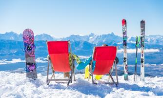 Two lounge chairs on a snow-covered mountain peak with a magnificent view of the mountains. A snowboard and skis are standing next to the lounge chairs.