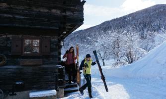 Two people are leaving a wooden cabin in the snow, carrying skis. Snow-covered mountains are visible in the background.
