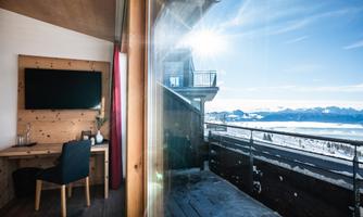 A cozy room with a desk and a television that overlooks a breathtaking mountain landscape. The sun shines through the windows, creating a bright atmosphere.