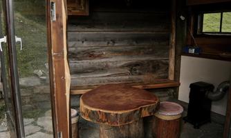 A rustic interior with a wooden bench and a round table made from a tree trunk. The walls are made of wood, and there is a small stove in the corner.