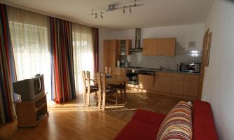 A cozy living room with a red sofa, a table, and chairs. The kitchen is modern and well-equipped, with large windows for plenty of daylight.