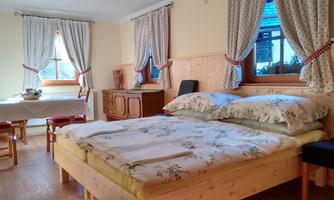 A cozy bedroom with a large bed and floral bedding. Windows with curtains and a table with chairs create an inviting atmosphere.
