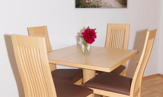 A bright dining area with a square table and four chairs. A vase with red flowers is on the table.