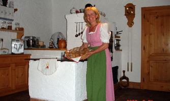 A woman in traditional attire holds fresh bread in a cozy kitchen. The surroundings are rustic with wooden cabinets and kitchen utensils.
