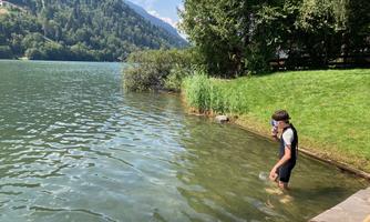 A boy stands in the clear water of a lake, surrounded by green trees and mountains. The sun is shining, creating a relaxed atmosphere.