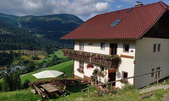 A cozy house with a red roof and beautiful flower balconies. The surrounding landscape features green hills and forests under a blue sky.