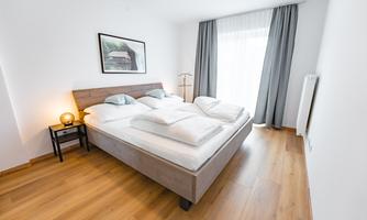 A modern bedroom with a large double bed and bright wooden floors. Curtains and a window provide plenty of daylight.