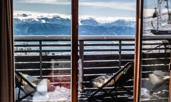 A bedroom with large windows that offer a view of snow-covered mountains. Outside, there are loungers on a balcony.