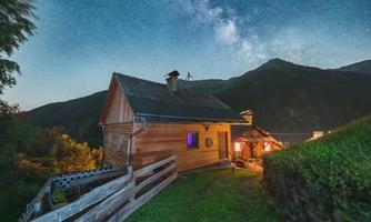 A cozy wooden house under a clear starry sky. In the background, mountains can be seen, and the building is softly lit.