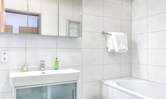 A modern bathroom with white tiles and a large mirror. It includes a sink area, a bathtub, and a green rug.