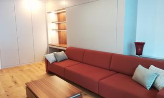A modern living room with an orange sofa and a wooden table. The walls are light and there are some decorative shelves in the background.