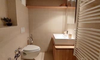 A modern bathroom with a sink, a toilet, and a bidet. There are wooden shelves and bright lighting.