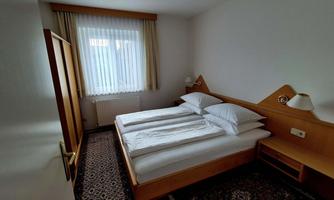 A simple bedroom with two single beds and wooden furniture. The room has a window with curtains and a small table.