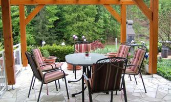 A cozy garden area with a table and chairs under a wooden overhang. Surrounded by green plants and a tranquil atmosphere.