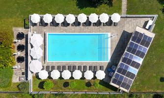 A swimming pool surrounded by loungers and sun umbrellas in a well-maintained garden. Solar panels are installed on the building next to the pool.
