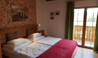 A cozy bedroom with two beds and a red blanket. The room has a balcony with a view outside and a blooming wall decoration.