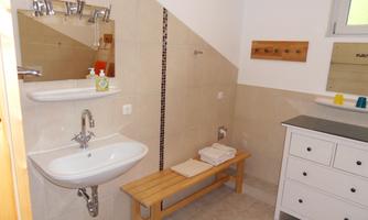 A modern bathroom with a sink, wooden bench, and a large mirror. The walls are tiled in light colors, and there is ample light.