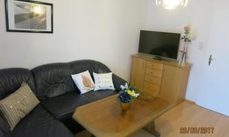 A cozy living room with a black sofa and a wooden table. In the background, there is a television and a sideboard with decorative plants.