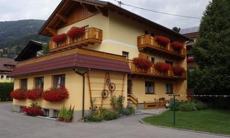 A charming two-story house with colorful flower boxes. It is surrounded by green plants and is situated in a mountainous area.