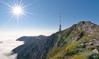 A mountain peak with a transmission mast and radiant sunshine. Below, clouds are visible surrounding the landscape.