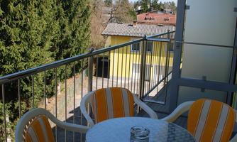 A balcony with two orange and white chairs and a round table. In the background, trees and additional buildings can be seen.
