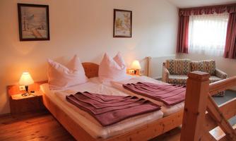 A cozy bedroom with two beds and pink blankets. There is a sofa and a window view with soft light.