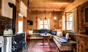 A cozy living space in a wooden cabin with a bench, table, and a wood stove. Large windows provide bright light and an inviting atmosphere.