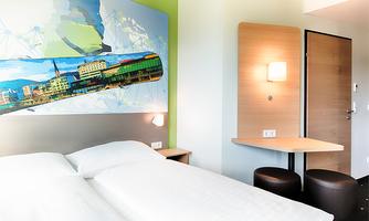 A modern hotel room with a double bed and a large wall mural. There is a table and chairs in the corner as well as a door that leads outside.