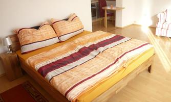 A cozy bedroom with a large bed and colorful cushions. In the background, a table and chairs are visible.