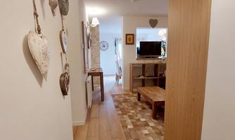 A bright corridor with wooden flooring and modern furniture. Decorative elements hang on the wall, and at the end of the corridor, a cozy living area is visible.
