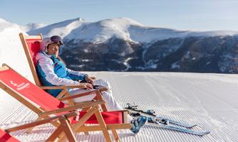 A woman is sitting relaxed on a deck chair in the mountains. In the background, there are snow-capped mountains and a clear blue sky.