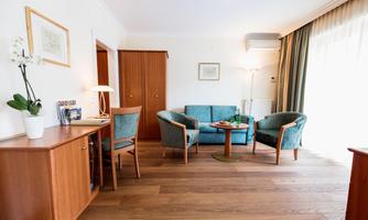 A cozy living space with two blue armchairs and a small table. The room has wooden floors and large windows that let in plenty of daylight.