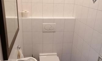 A small, clean bathroom with white tiles. It includes a sink, a toilet, and a mirror.