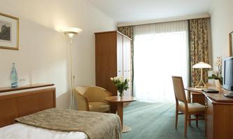 A cozy hotel room with a bed, a table, and a chair. Large windows let in a lot of natural light and offer a view of the terrace.