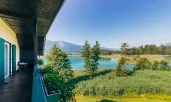 A view from the balcony of a clear lake with lush greenery and mountains in the background. The sky is blue and cloudless.