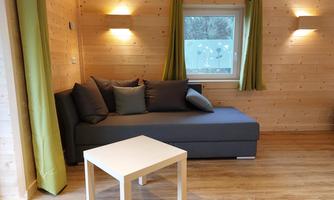 A cozy living space with a gray sofa and a white table. The walls are made of wood and there are green curtains at the windows.