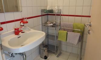 A modern bathroom with white tiles and a red accent. On a shelf, there are personal care products and fresh towels in various colors.