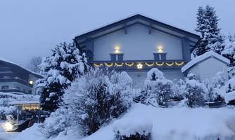 A snow-covered house at dusk, surrounded by snow-covered trees. The lights on the balcony create a festive atmosphere.