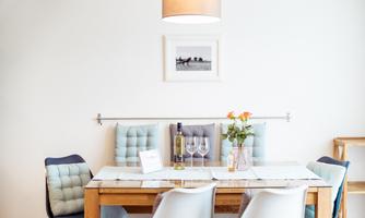 An inviting dining area with a wooden table and light chairs. On the table, there is a vase with flowers and a bottle of wine.