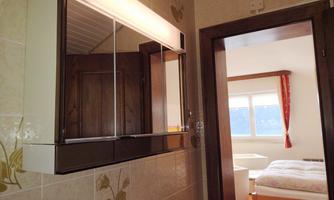 A bathroom with a mirror and a lighted cabinet. In the background, there is a bedroom with a bed and windows.