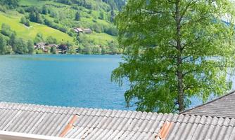 A cozy balcony overlooking a clear lake and lush green hills. The landscape is framed by mountains and a blue sky.