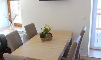 A bright dining area with a wooden table and six chairs. A television hangs on the wall, and there is a door that leads to a balcony.
