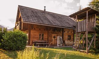 A cozy wooden house with a rustic roof and a small veranda. Next to it is a woodpile and a play tower in the garden.
