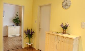 A bright entrance area with yellow walls and a wooden cabinet. Fresh flowers and a wall clock are on the shelf.