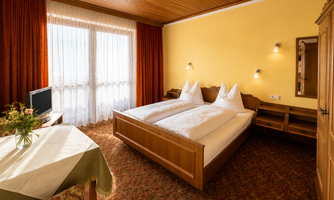 A cozy bedroom with a double bed and bright walls. The decor is simple and inviting, with a view of daylight through large windows.