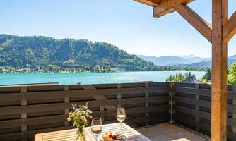 A sunny terrace overlooking a picturesque lake and the surrounding mountains. On the table, there is a bouquet of flowers and two glasses, accompanied by a plate of fresh fruit.